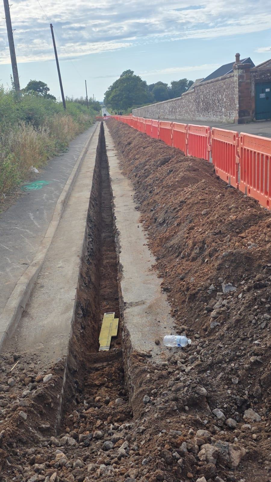 Long straight trench along a footway with spoil heaped behind barriers