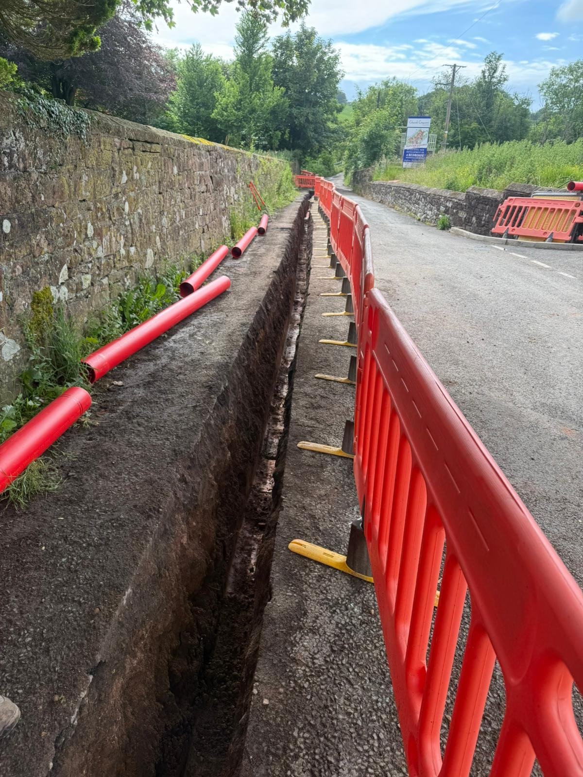 Open trench along a sandstone boundary wall with red duct lengths laid out ready
